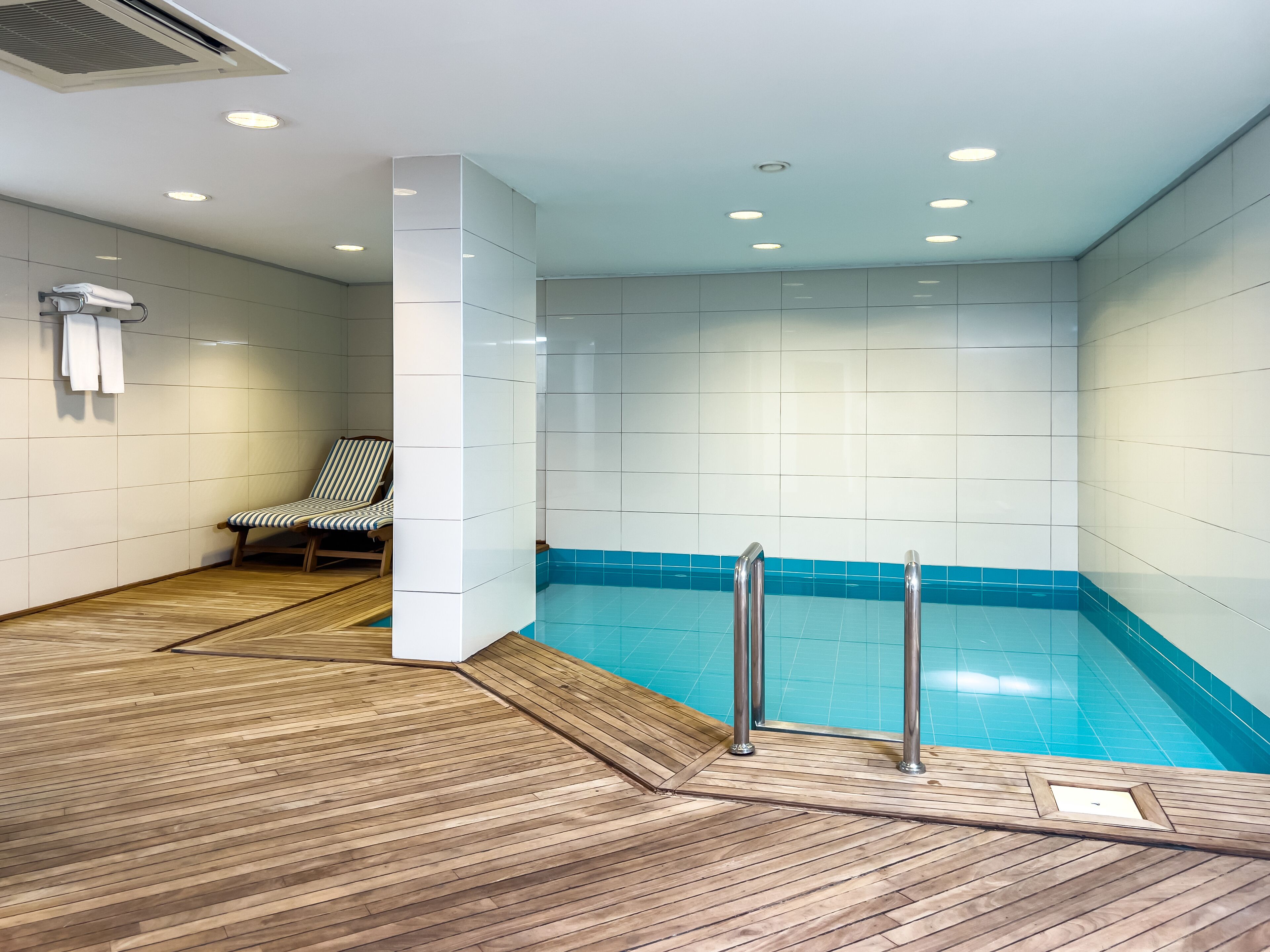 Una piscina interior moderna con una zona de terraza de madera, una tumbona y un perchero para toallas.
