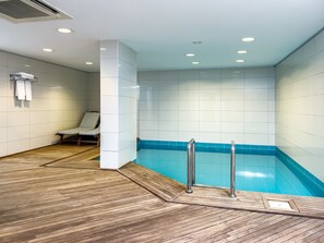 Una piscina interior moderna con una zona de terraza de madera, una tumbona y un perchero para toallas.