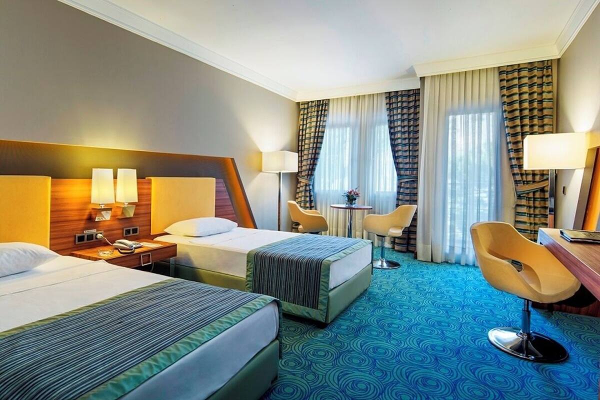 A hotel room with two beds, a desk, a chair, a small table, and a lamp.