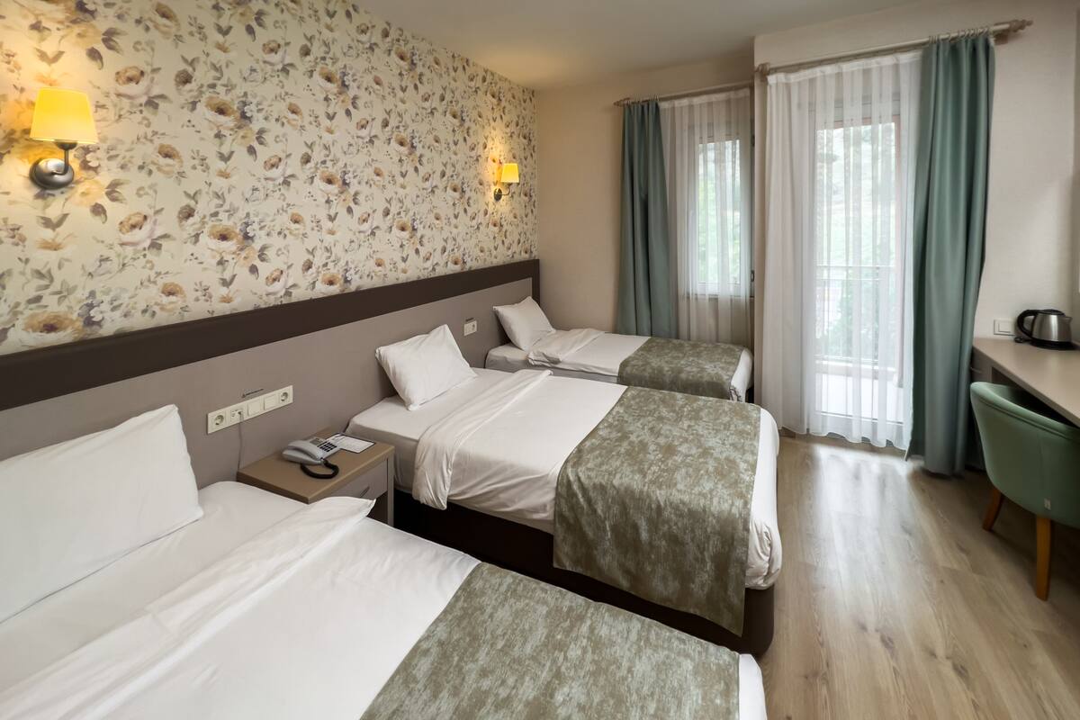 A hotel room with two beds, a desk, and a chair. There are two bedside tables with lamps and a telephone. The room has floral wallpaper and a window with curtains.