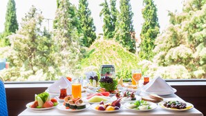 Une table de petit-déjeuner garnie de divers fruits, légumes et boissons, le tout mis en valeur par un décor de grands arbres et un ciel dégagé.