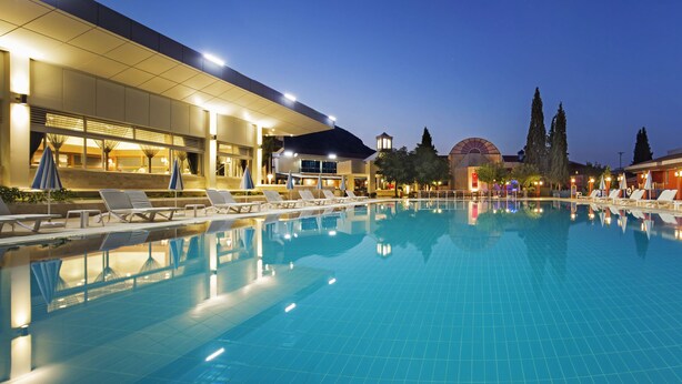Une piscine entourée de bâtiments à l’éclairage chaleureux, avec des chaises longues et des parasols.