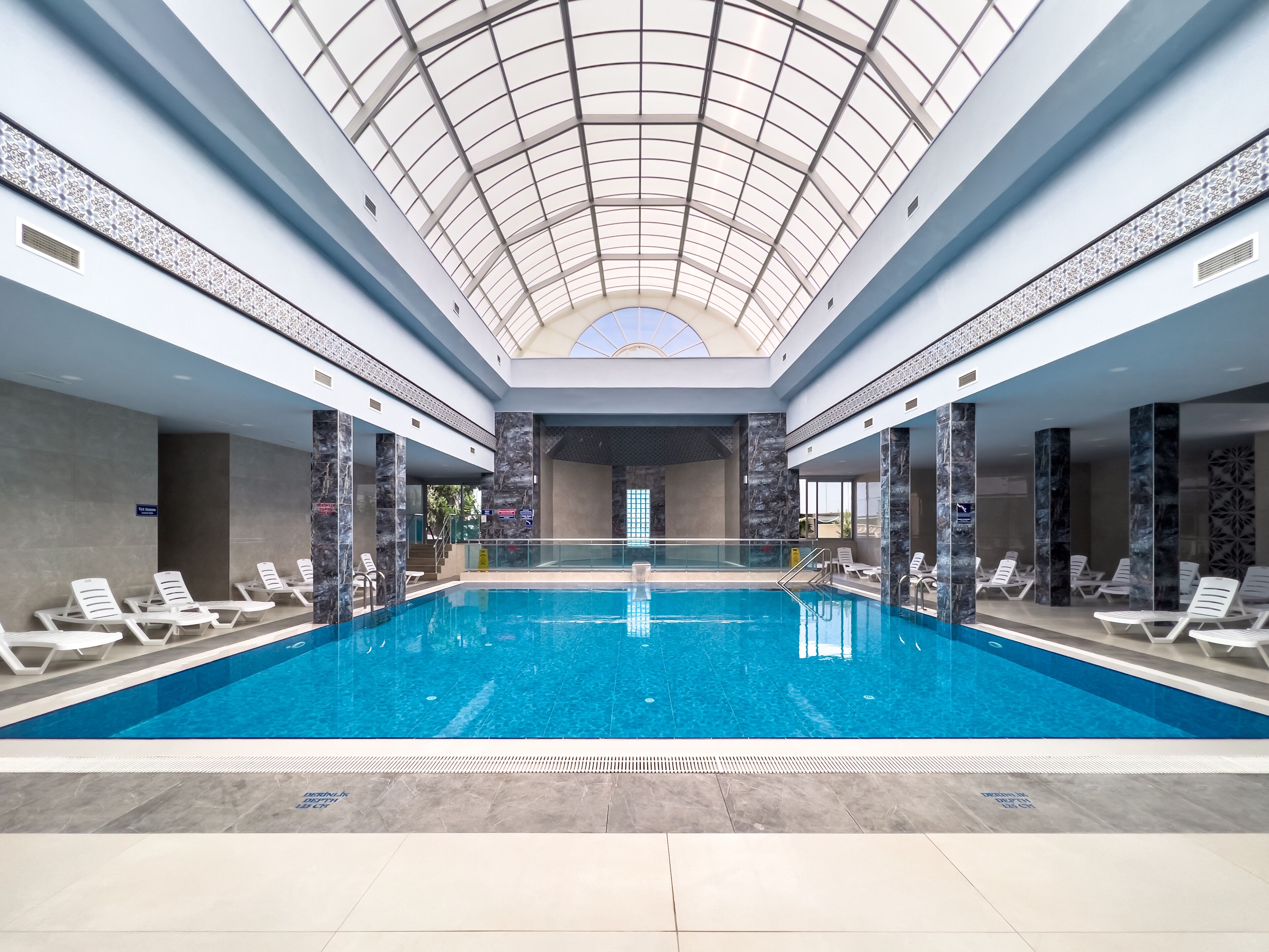 An indoor swimming pool with a glass ceiling, surrounded by lounge chairs and marble walls.