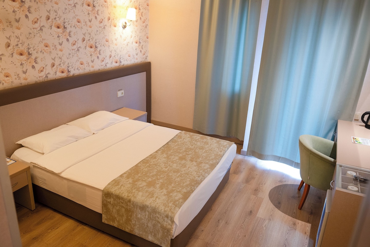 A hotel room with a bed, a nightstand, a desk with a kettle, and a chair.