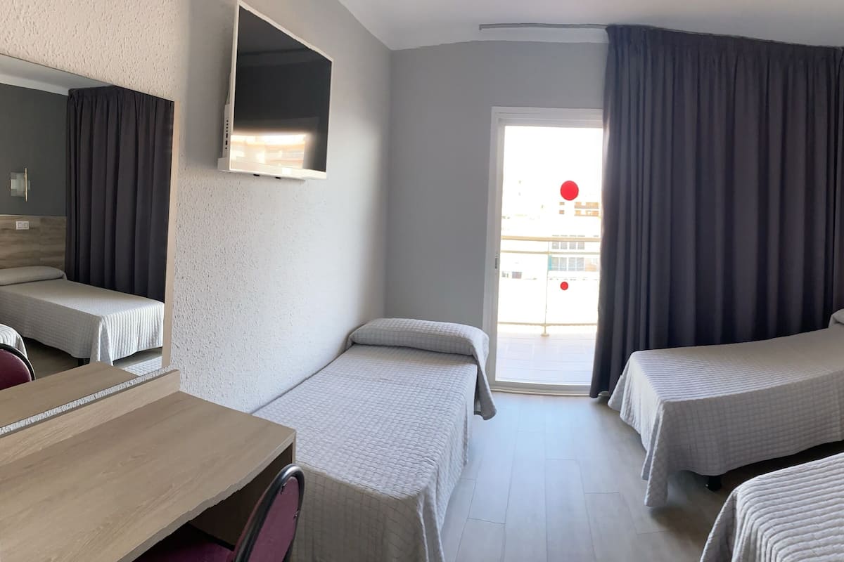 Habitación de hotel con tres camas individuales, un escritorio de madera, una mesita pequeña, un espejo, un televisor y una ventana con cortinas.