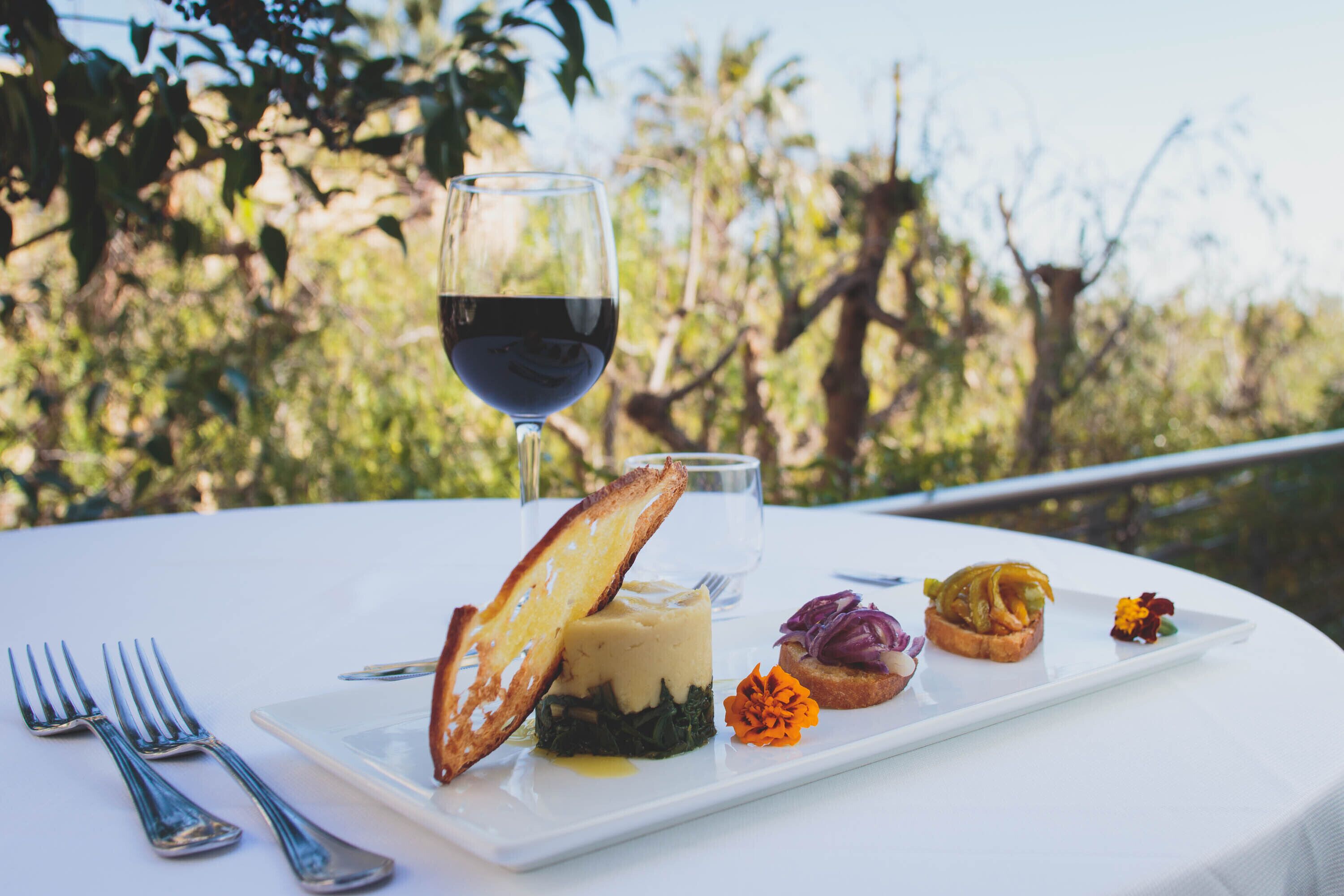A white table with a plate of food, a glass of red wine, and a fork and knife on a white tablecloth.