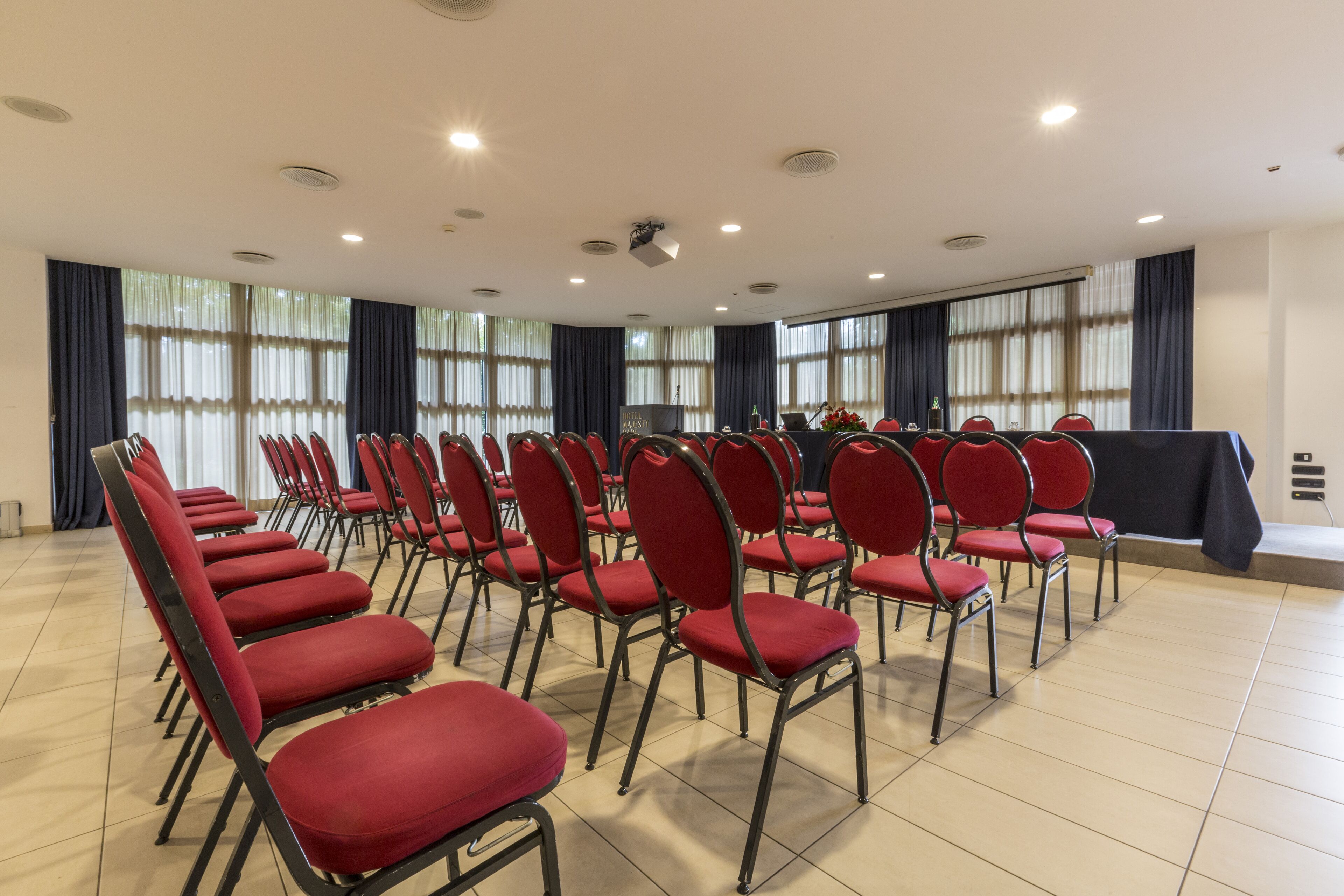 Una sala conferenze con file di sedie rosse, un podio e ampie finestre con tende.