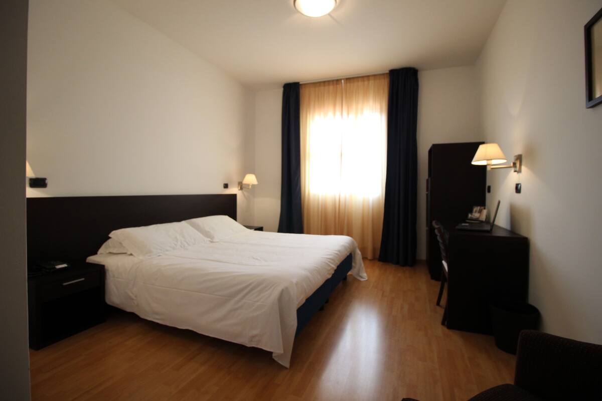 Une chambre d’hôtel avec un lit, un bureau, une chaise, une lampe et une fenêtre avec des rideaux.