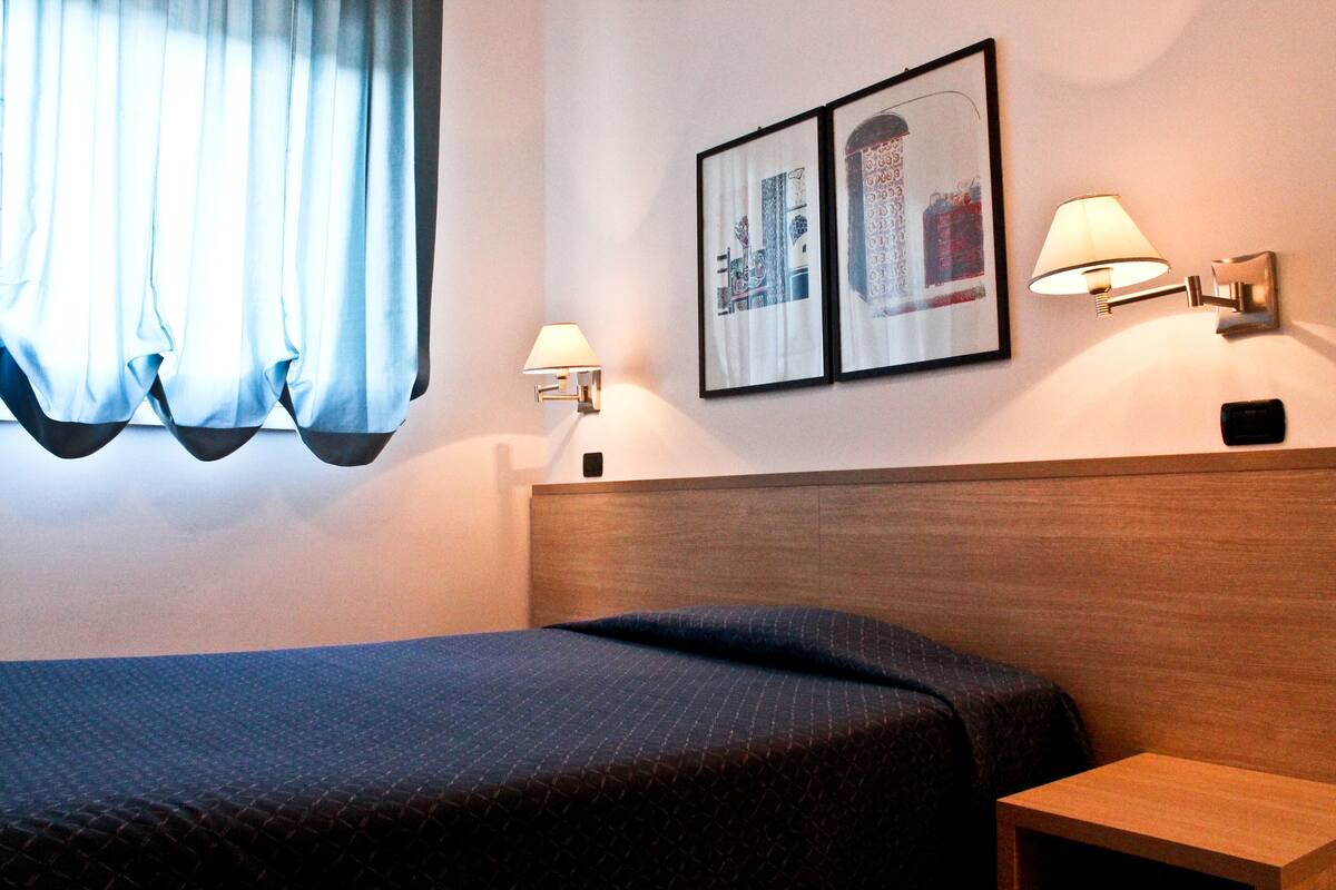 Une chambre d’hôtel avec un lit, deux lampes fixées au mur et un tableau encadré accroché au mur.