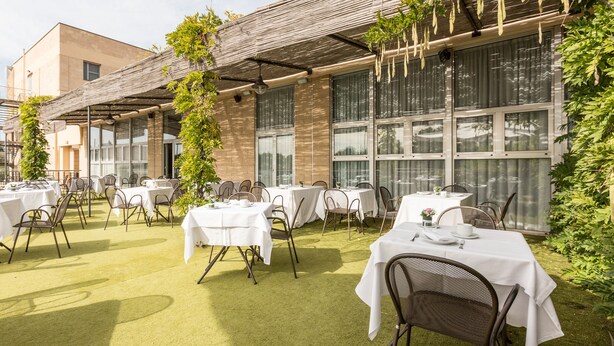 Un área de comedor al aire libre con mesas y sillas, un espacio de estar cubierto y un edificio con ventanas amplias y techo de madera.