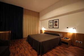 A hotel room with a wooden floor, a bed with a brown bedspread, a nightstand with a lamp, and two framed pictures on the wall. - Hotel Majesty (Bari)