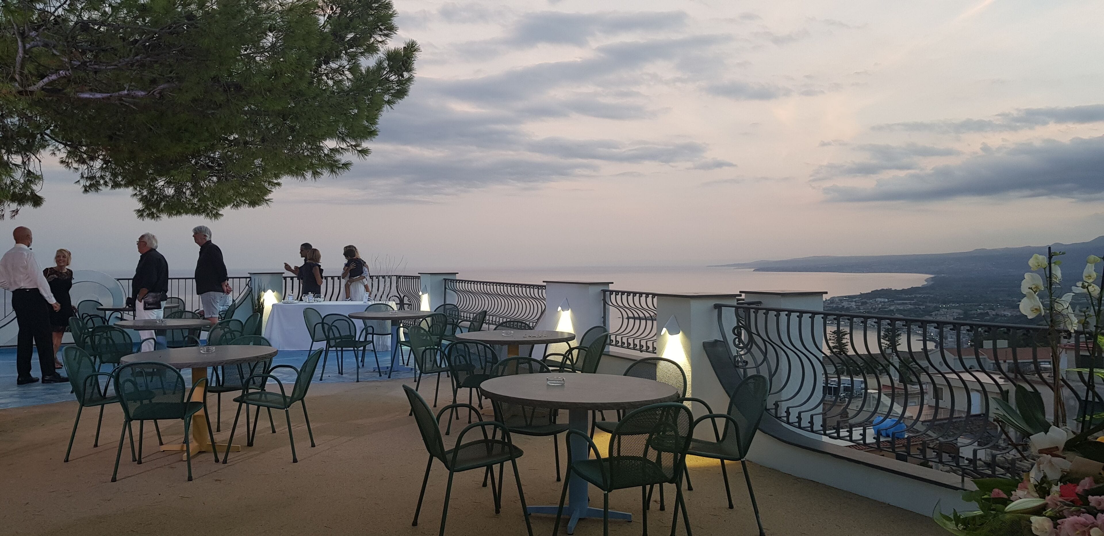 An outdoor terrace with round tables and chairs, overlooking a scenic view of the sea and distant hills.