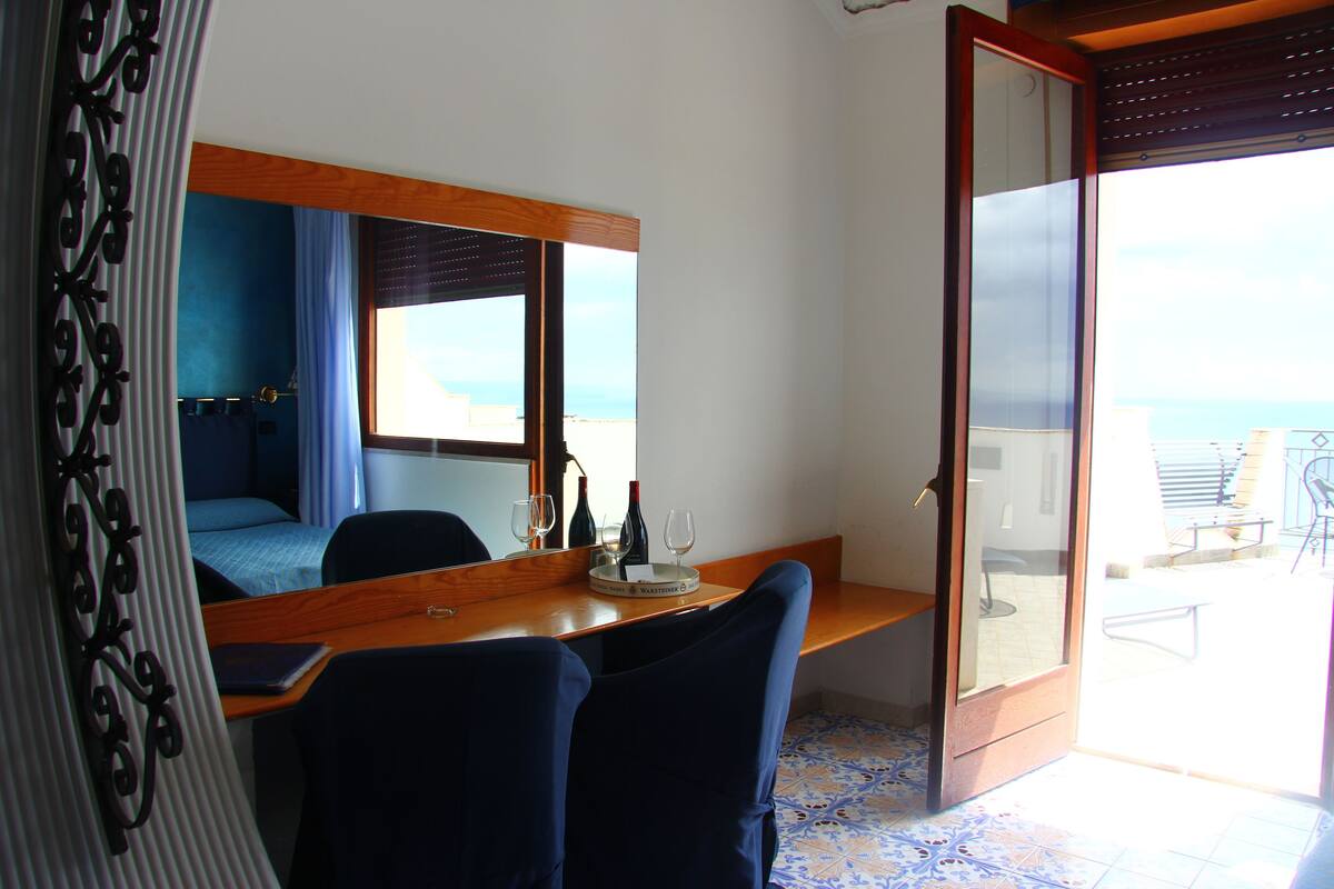 A room with a bed, a desk with a mirror, a chair, and a view of the sea through a large window.