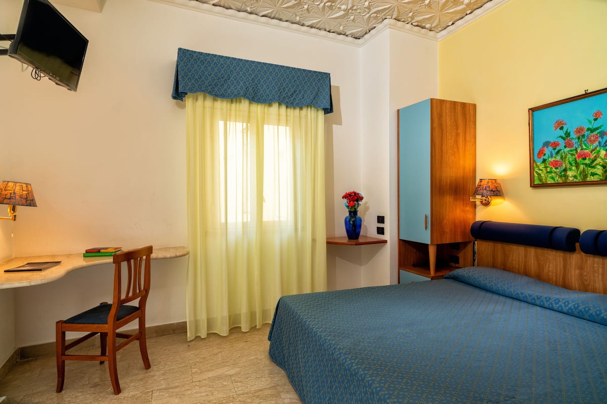 A hotel room with a bed, a desk, a chair, a TV, a painting, and a window with curtains.