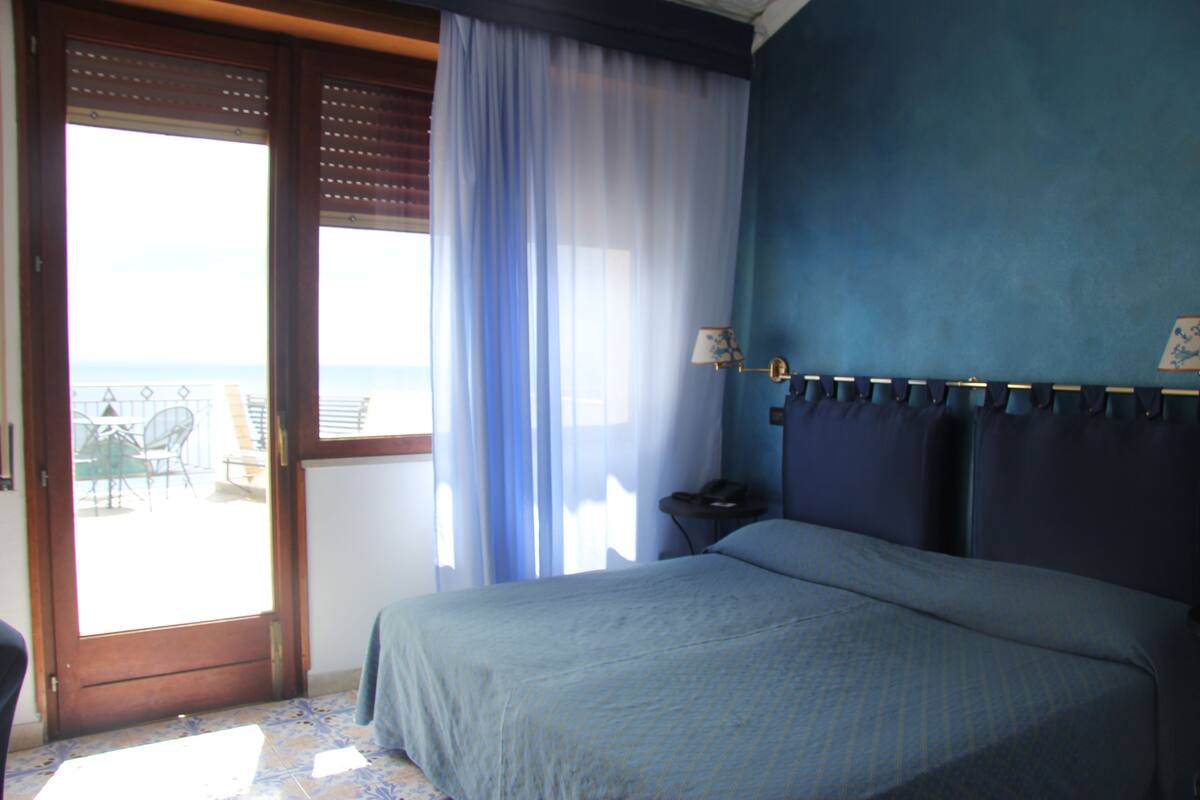 A bedroom with a bed, a nightstand, a lamp, and a view of the outdoors through a door.