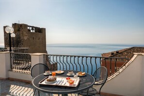 A balcony with a table set for breakfast, overlooking a coastal view and a historic building. - Hotel Vello d'Oro (Taormina)