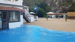 A pool area with a blue floor, a building with a sign reading "Cafikano Makanou," outdoor seating, and a rocky backdrop. - Hotel Vello d'Oro (Taormina)