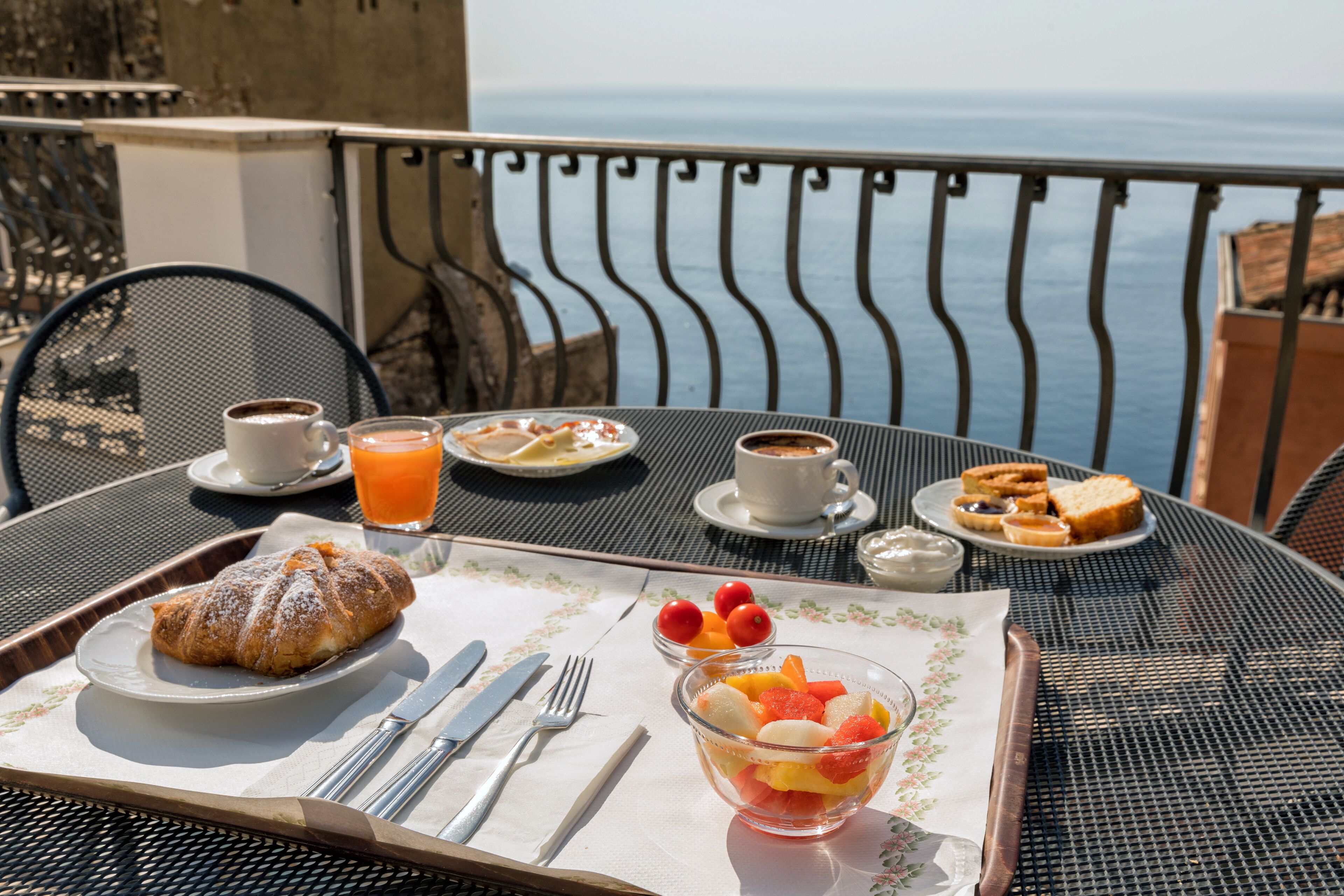Ontbijt met croissant, fruitmand, koffie en sap op een balkon met uitzicht op zee.