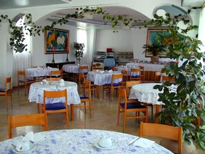 A dining area with round tables covered with floral tablecloths, wooden chairs, and potted plants. - Hotel Vello d'Oro (Taormina)