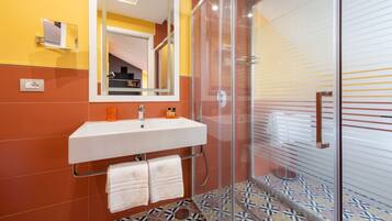 A bathroom with a white sink, orange and yellow walls, a glass shower enclosure, and patterned floor tiles.