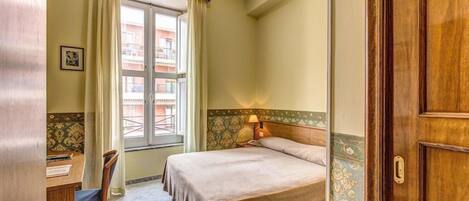 Una habitación de hotel con cama, escritorio con silla, ventana con cortinas y puerta.