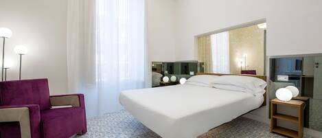 A modern hotel room with a large bed, a purple armchair, a bedside table with a lamp, and a large mirror.