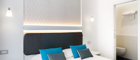Quarto de hotel moderno com cama bem arrumada, mesa de cabeceira com telefone e banheiro visível por uma porta aberta.