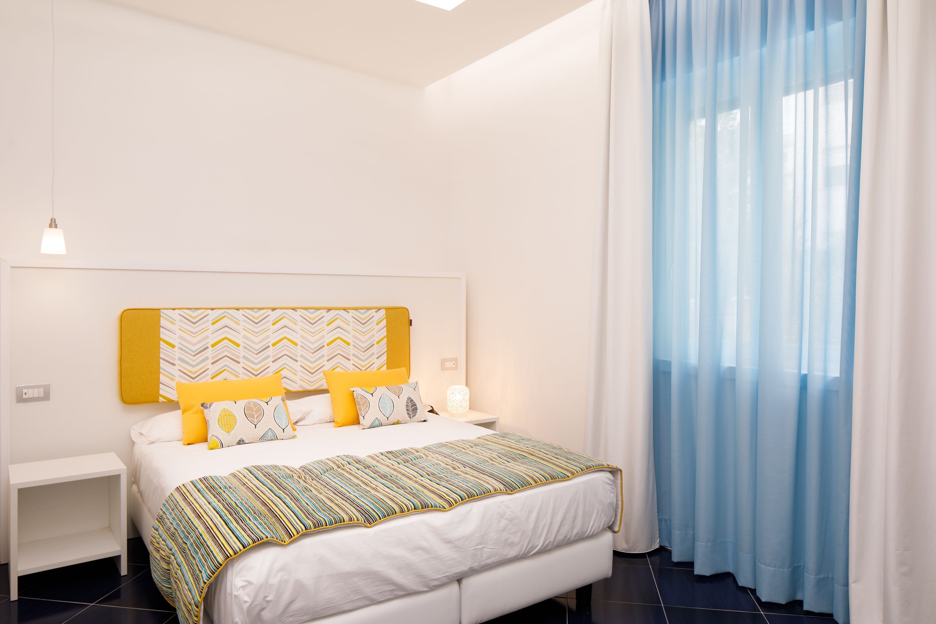 A modern bedroom with a bed, nightstands, a bedside table, and a window with sheer curtains.