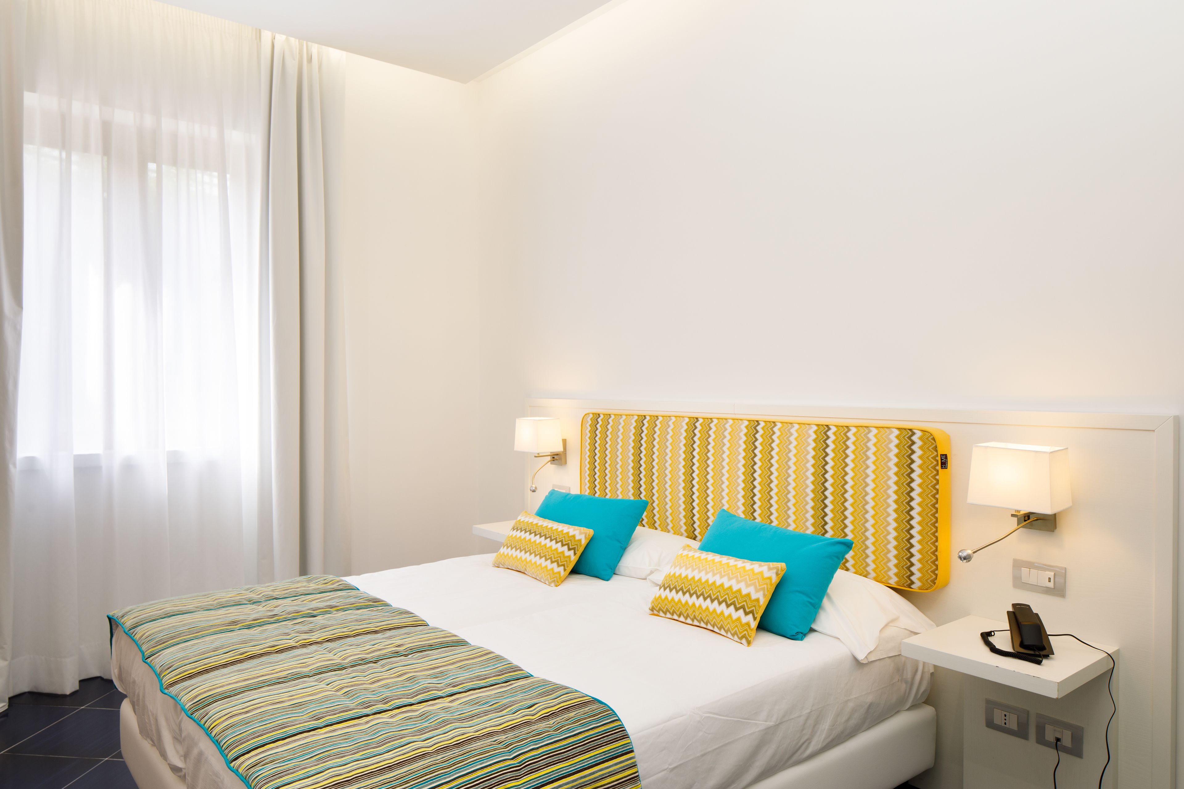 A hotel room with a bed, two bedside lamps, a telephone, and a window with curtains.