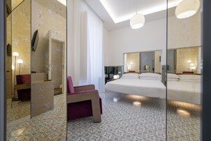 Una habitación de hotel moderna con una cama grande, una zona de estar con una silla, un tocador con espejo y un ventanal con cortinas.