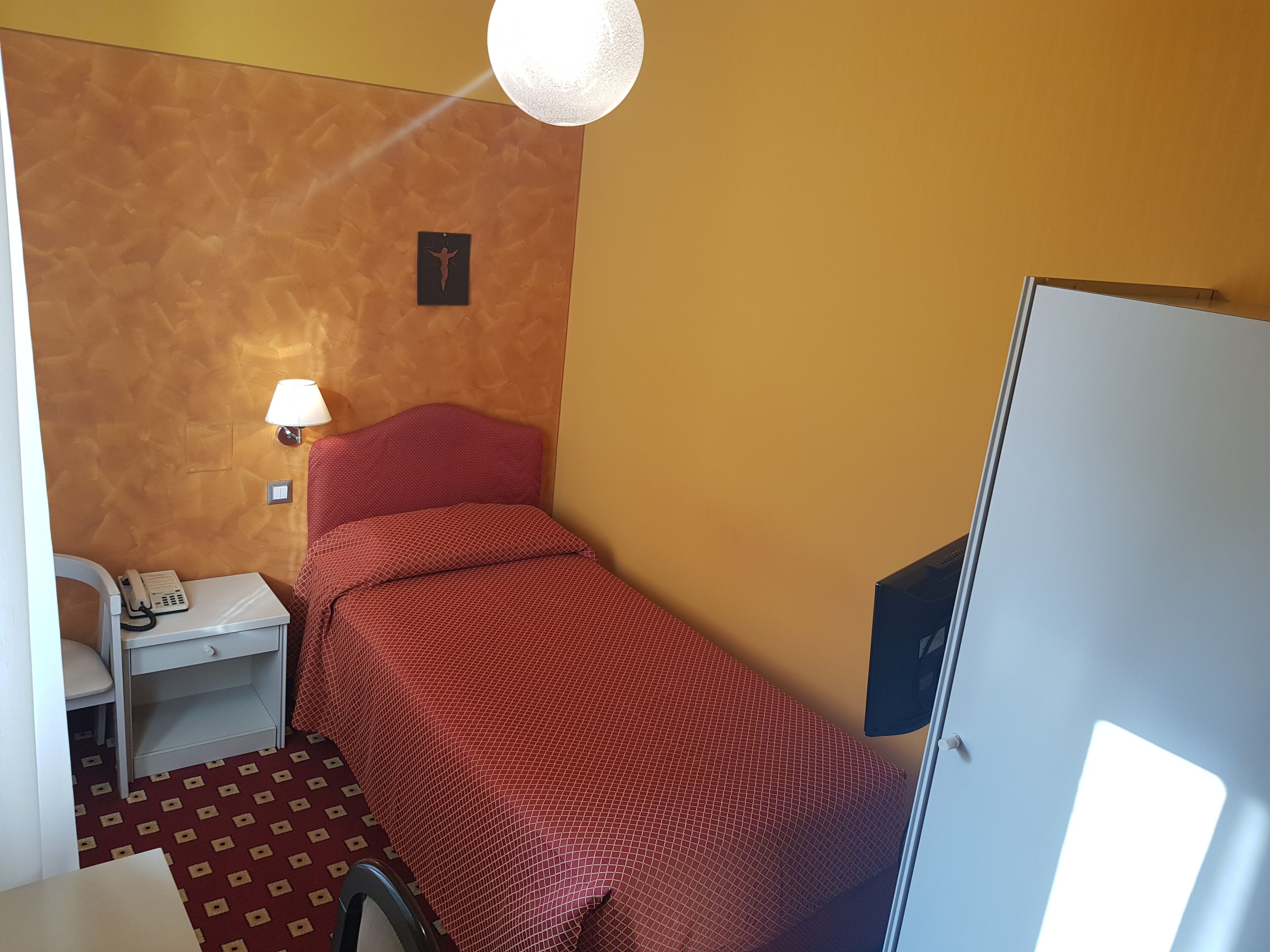 Una habitación pequeña con una cama individual, colcha roja, lámpara de noche, teléfono sobre una mesita y un armario.