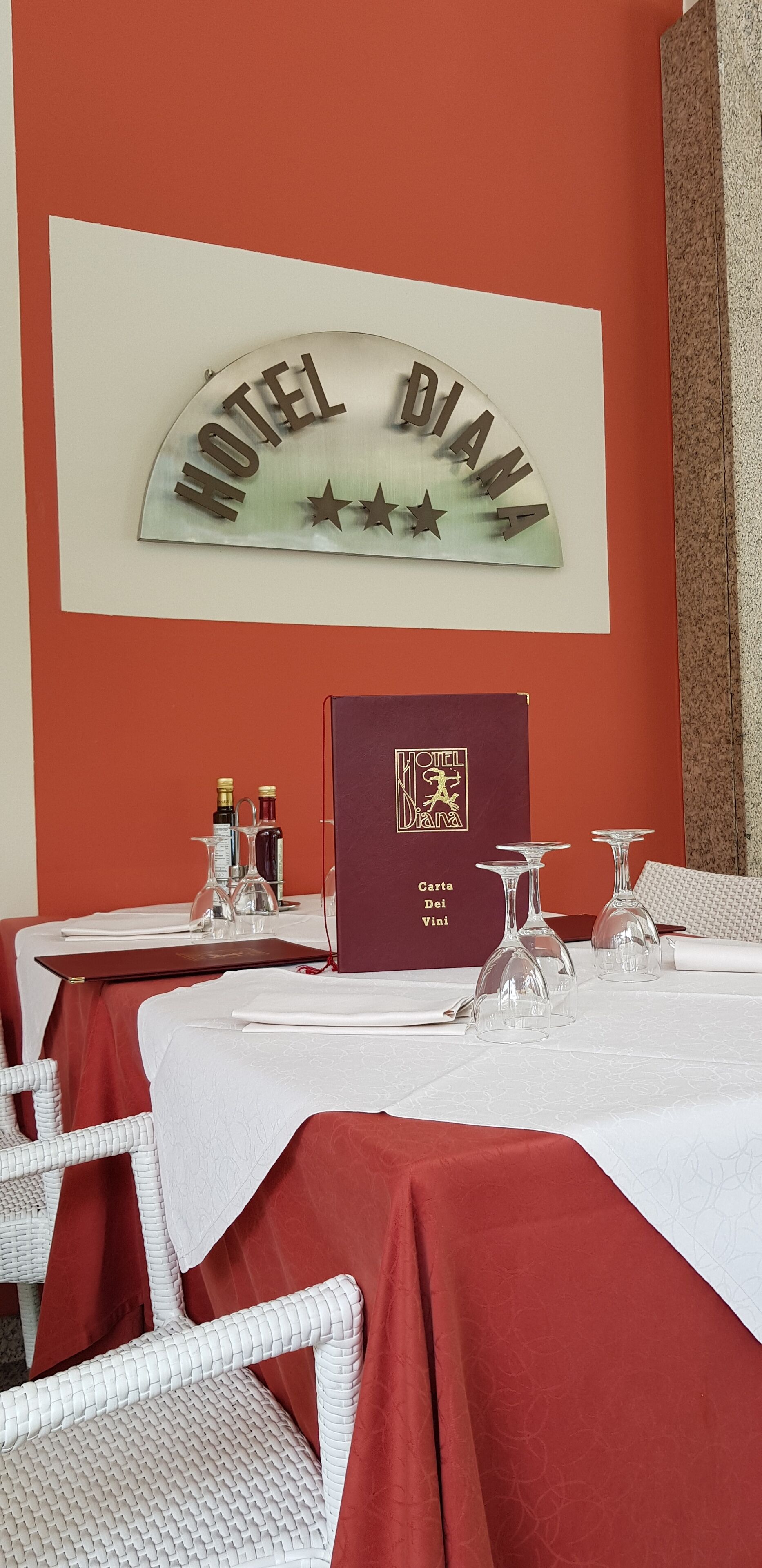 Une chambre d’hôtel aux couleurs rouge et blanc, avec une enseigne indiquant « HÔTEL DIANA » et un petit livre rouge intitulé « Carta Del Vini ».