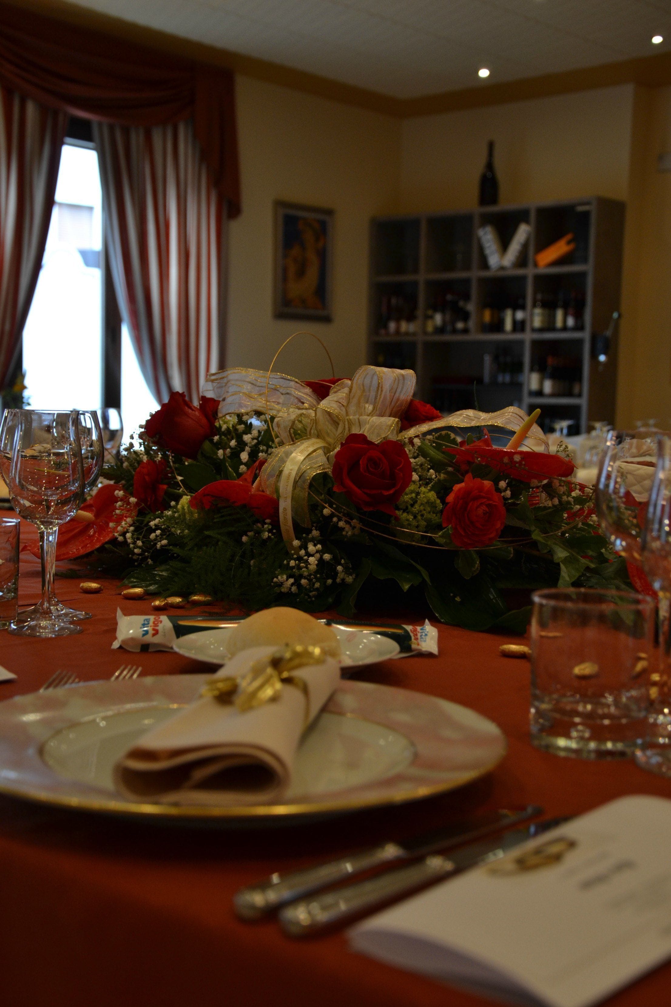 Una mesa de comedor dispuesta con rosas rojas, un ramo y una botella de vino en una habitación con una estantería y un cuadro en la pared.