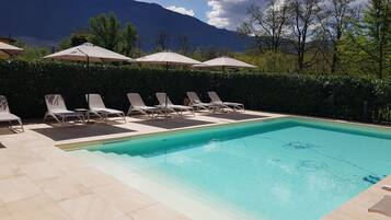 Une piscine entourée de chaises longues et de parasols, bordée d’une haie et avec des montagnes en toile de fond.