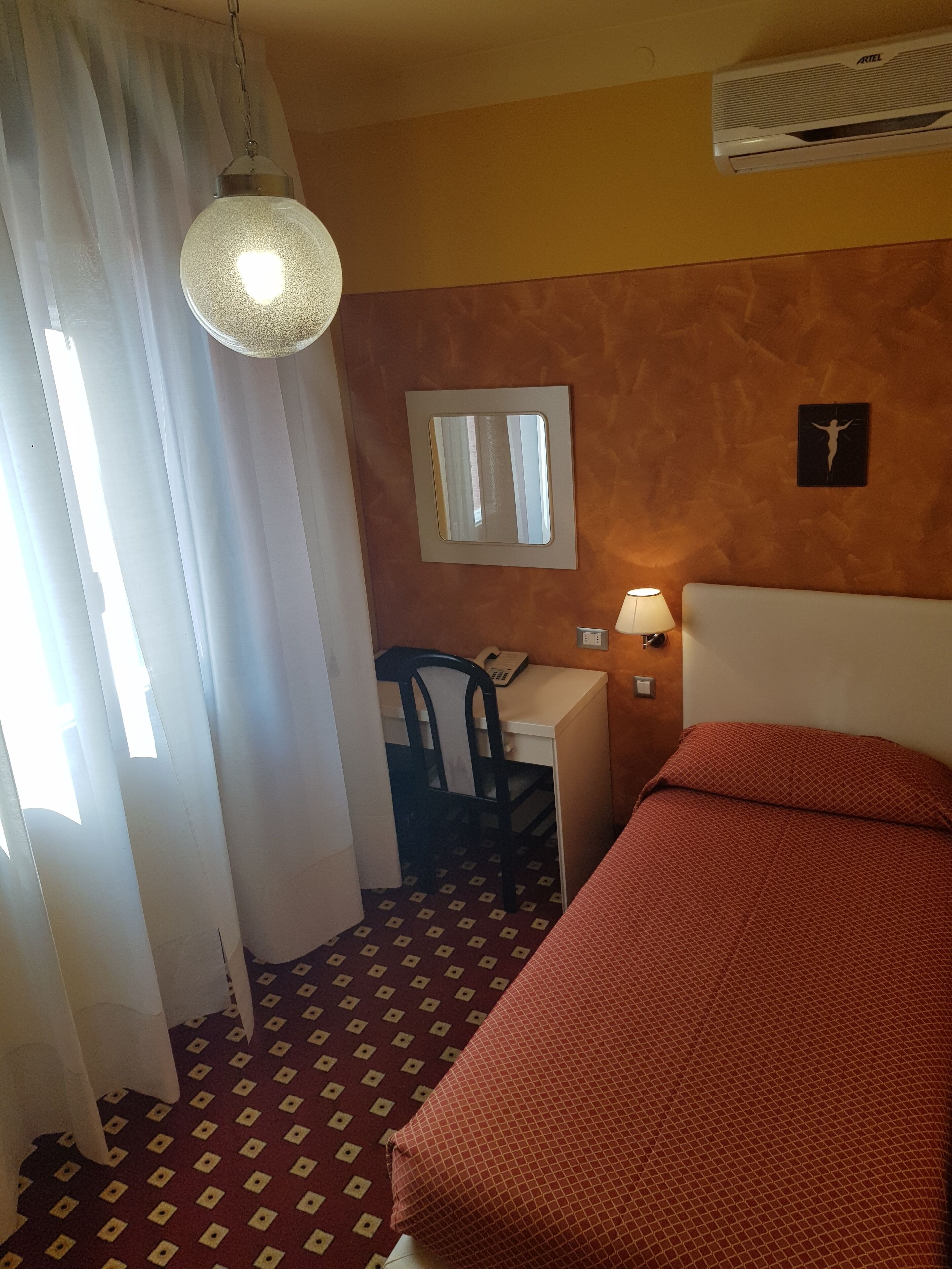 A hotel room with a bed, a desk with a chair, a hanging light fixture, a wall-mounted air conditioner, and a window with curtains.