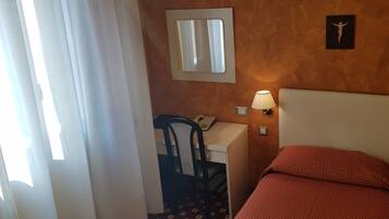 A hotel room with a bed, a desk with a chair, a hanging light fixture, a wall-mounted air conditioner, and a window with curtains.