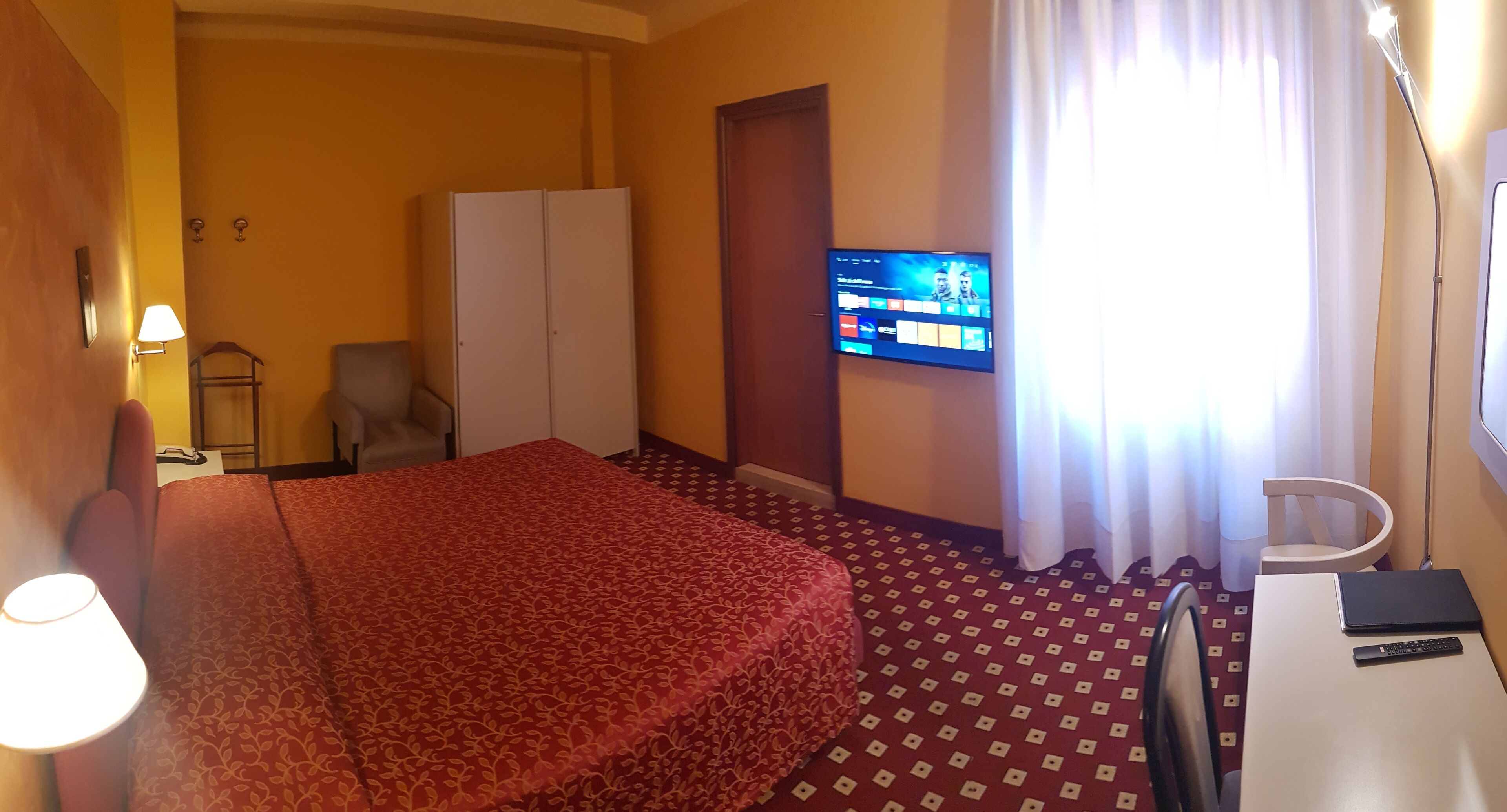 A hotel room with a bed, a desk with a laptop, a TV on the wall, and a wardrobe.