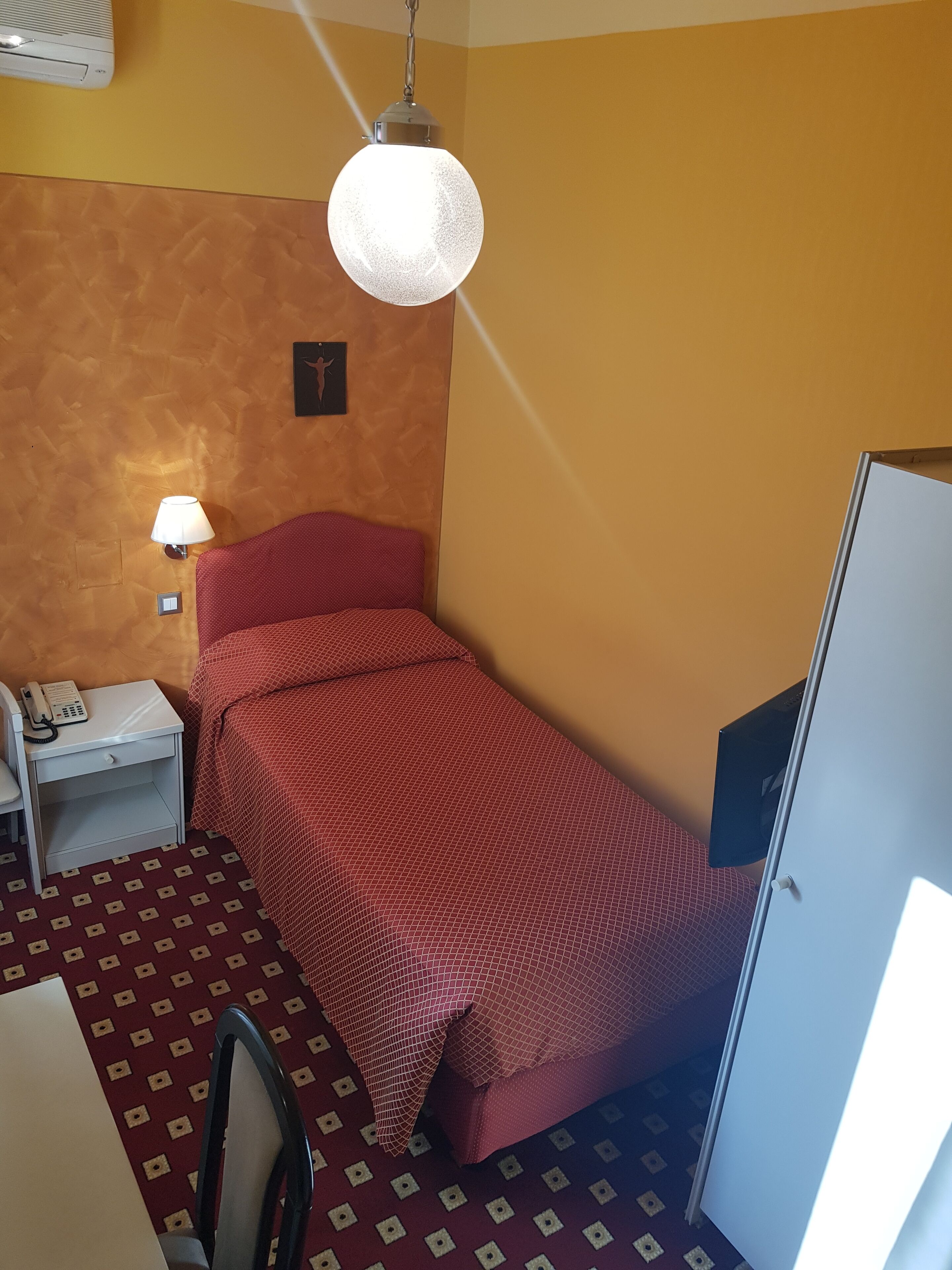 Une petite pièce, de taille modeste, comprenant un lit simple, un bureau avec une chaise, un téléphone et une lampe fixée au mur.