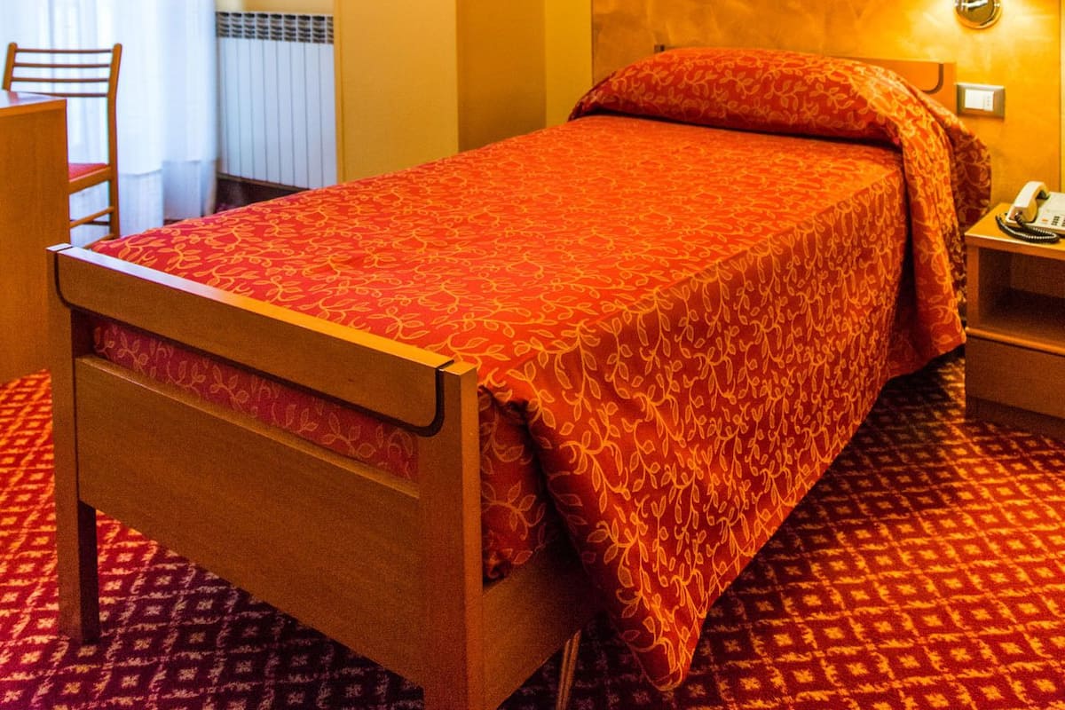 Une chambre d’hôtel avec un lit en bois, une couvre-lit rouge à motifs, une lampe blanche sur la table de chevet, une chaise et une fenêtre avec des rideaux.