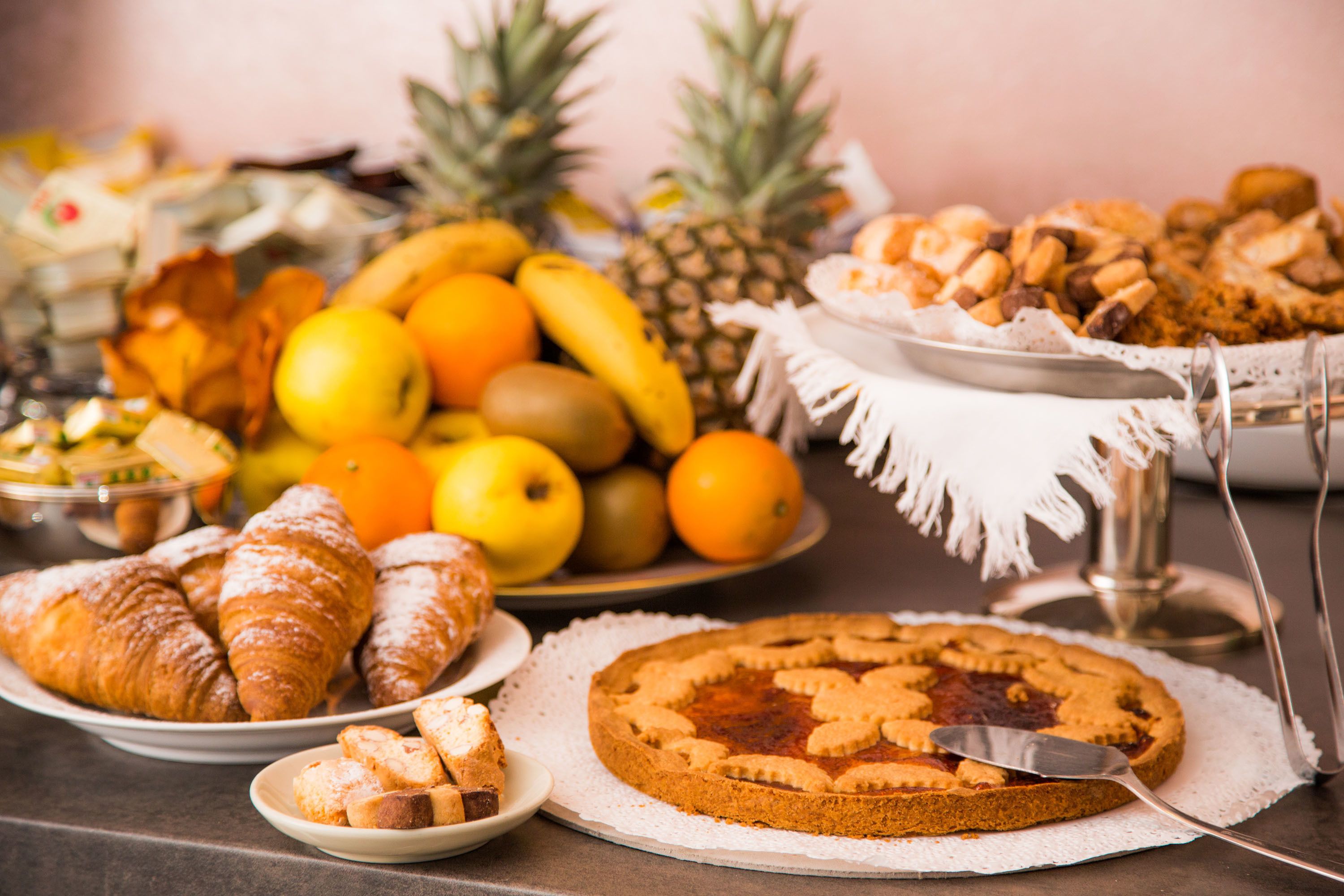 Verschiedene Backwaren, darunter Croissants, ein Kuchen und eine Auswahl an Gebäck, auf einem Tisch. Im Hintergrund sind Früchte wie Bananen und Orangen zu sehen.