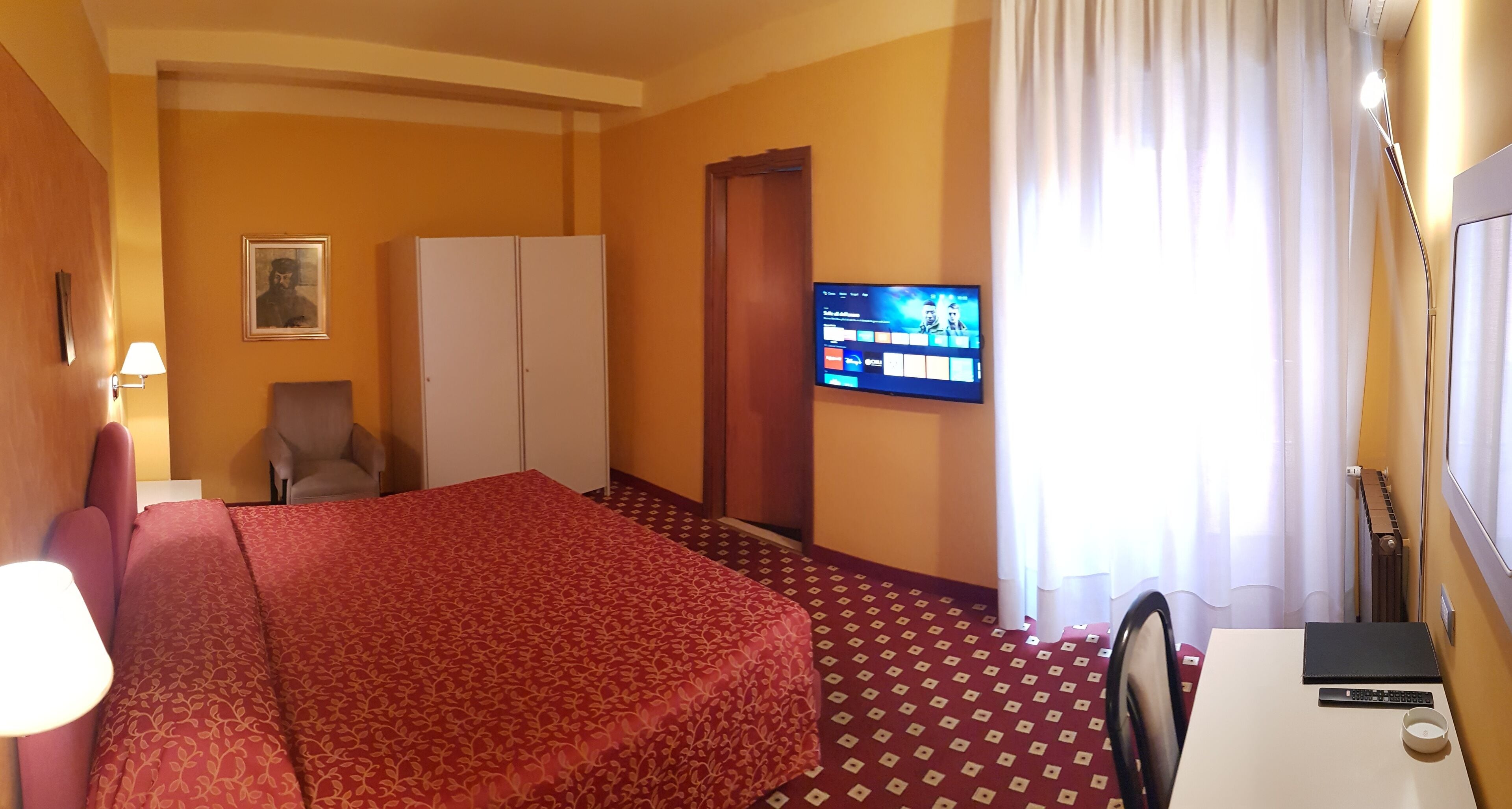 Una habitación de hotel con cama, una silla, escritorio, televisor y ventana con cortinas.