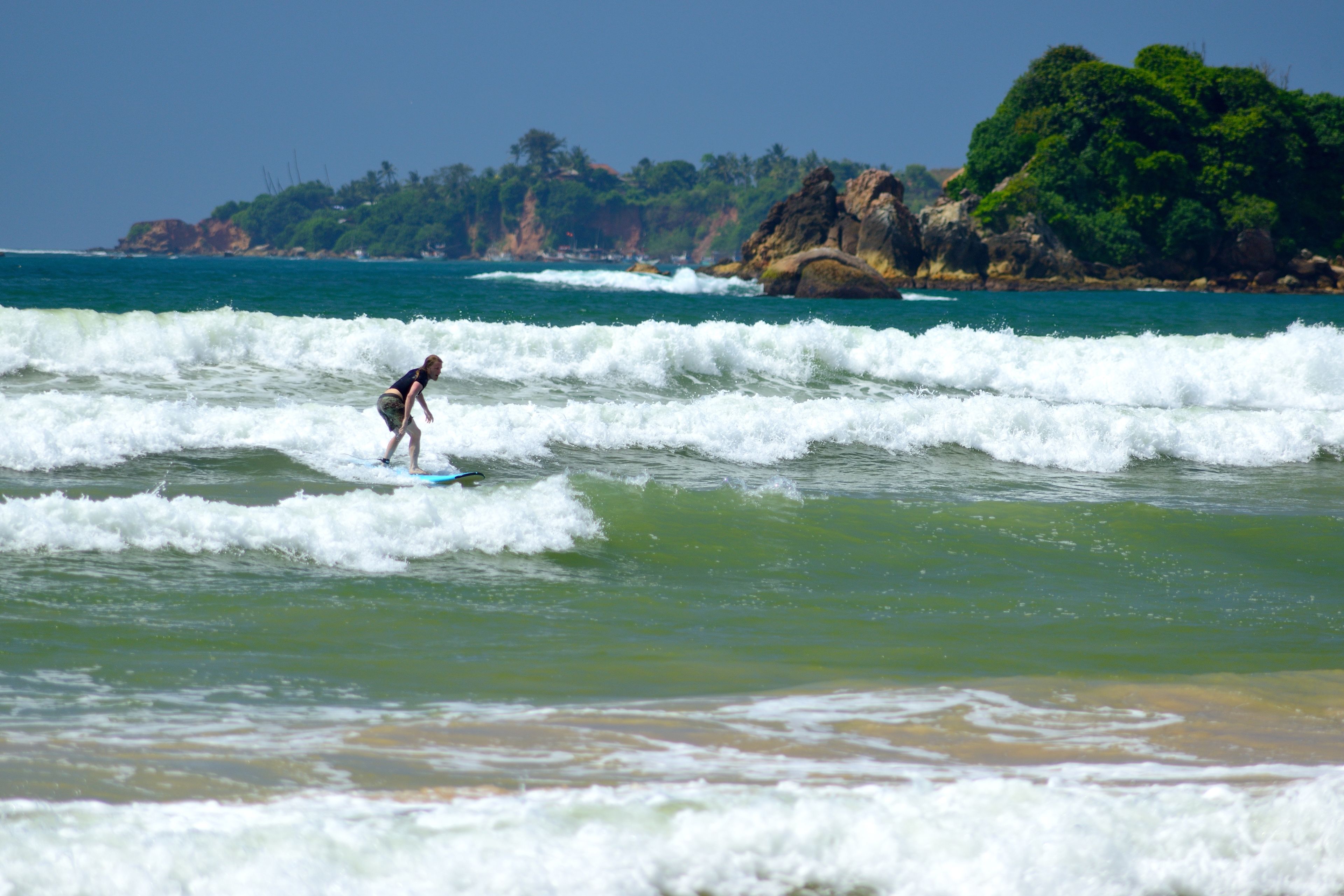 A beach with waves, a surfer, and a rocky coastline with greenery.