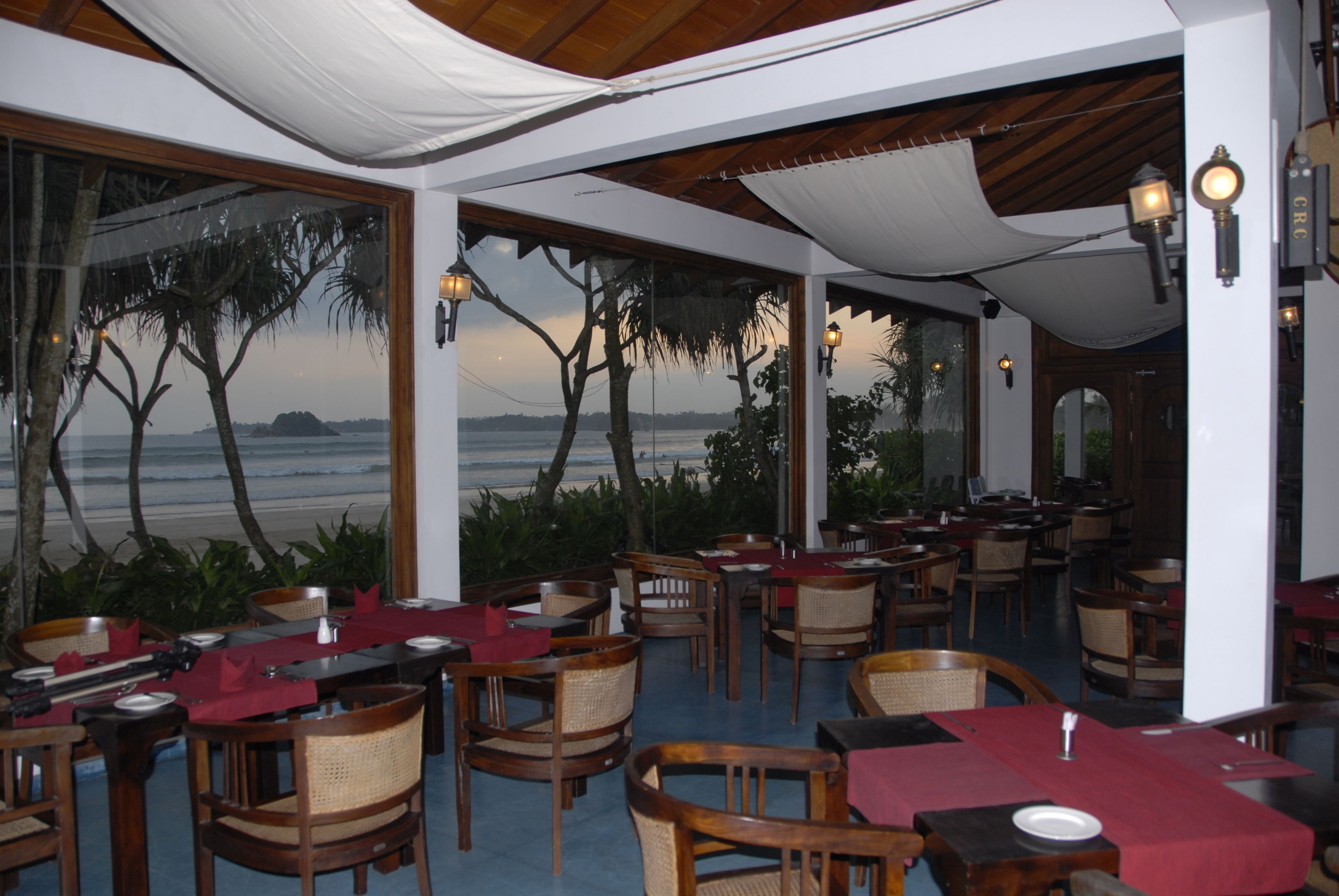 A restaurant with tables set for dining, large windows offering a view of the sea, and a covered outdoor area with wooden furniture.