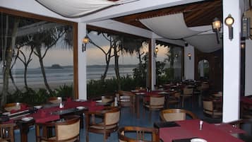 Un restaurant avec des tables dressées pour le service, de grandes fenêtres donnant sur la mer et un espace extérieur couvert aménagé avec du mobilier en bois.