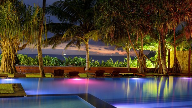 A swimming pool with a view of the ocean at dusk, surrounded by palm trees and lounge chairs. - Weligama Bay Resort (Weligama)