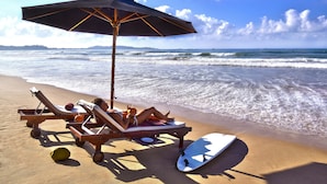 A beach scene with two lounge chairs under an umbrella, a surfboard, and a beach ball on the sand. - Weligama Bay Resort (Weligama)