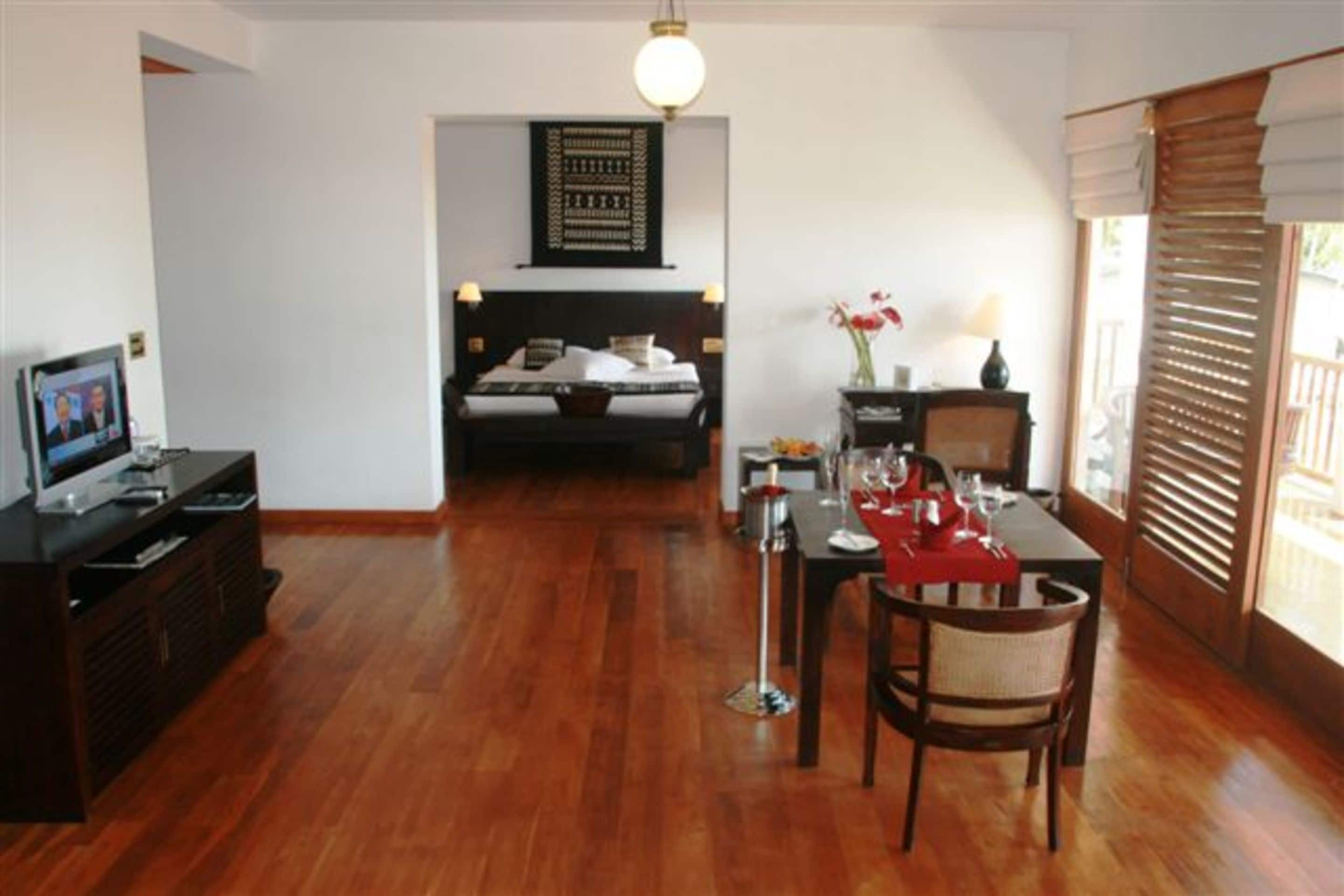 A hotel room with a wooden floor, a bed with a headboard, a dining table with chairs, a TV, and a window with blinds.