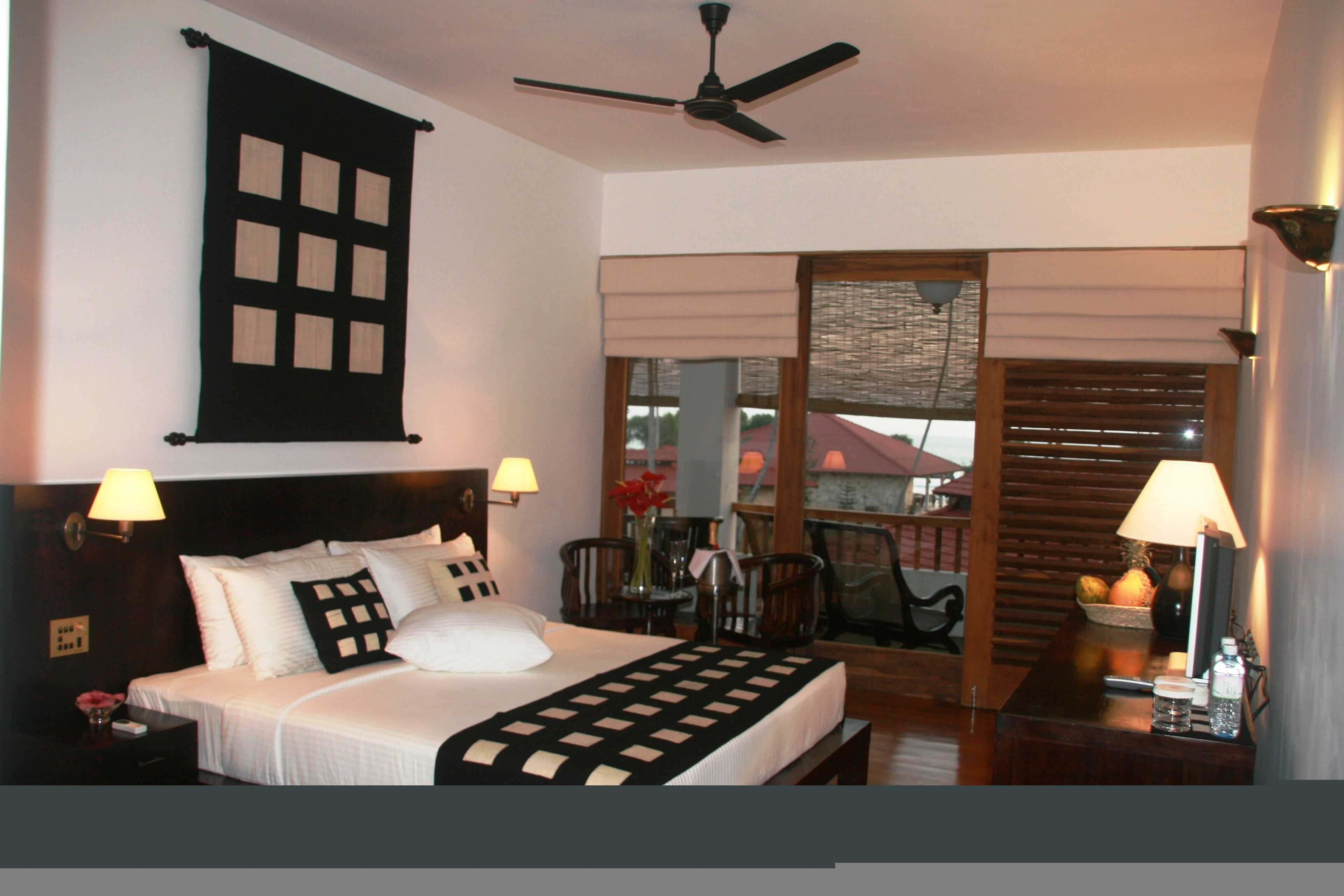 A hotel room with a bed, two bedside lamps, a ceiling fan, a window with blinds, a small table with a vase and fruit, and a view of a building outside.