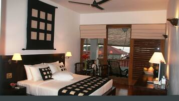 Habitación de hotel con cama, dos lámparas de noche, ventilador de techo, ventana con persianas, mesita con un florero y fruta, y vista a un edificio.