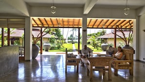 A spacious indoor area with a wooden bench and a table, large potted plants, and a view of a tropical garden. - Weligama Bay Resort (Weligama)